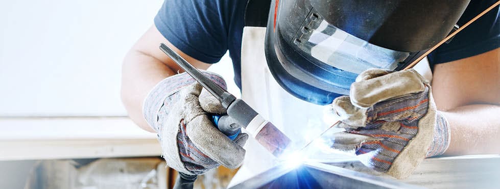 Male in face mask, protective gloves welds with argon-arc welding. Welder makes weld seam on metal frame. Worker dressed in t-shirt and overalls.