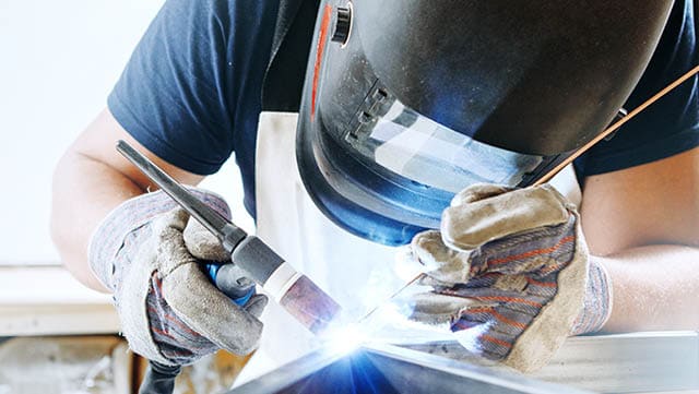 Male in face mask, protective gloves welds with argon-arc welding. Welder makes weld seam on metal frame. Worker dressed in t-shirt and overalls.