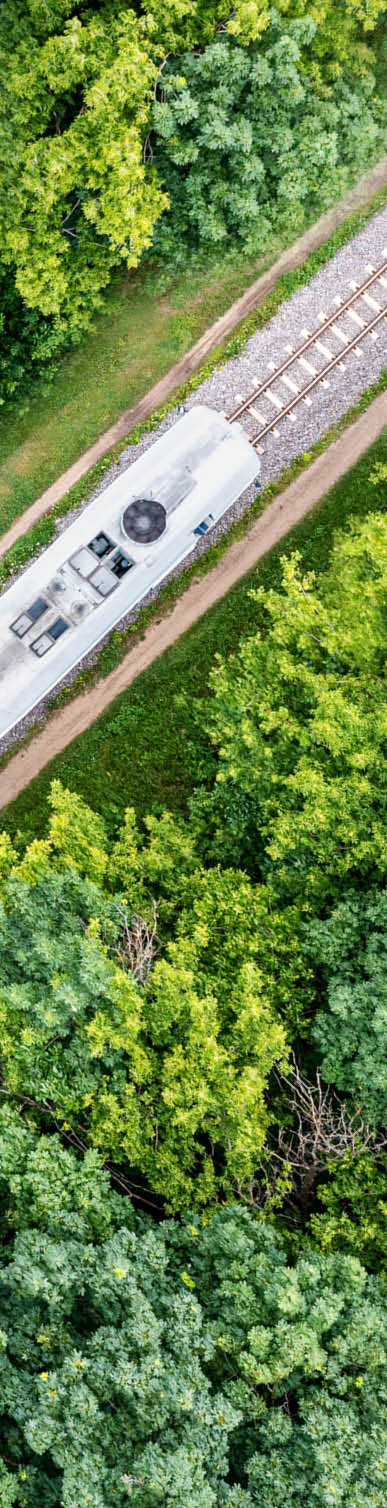 A diesel locomotive pulls the carriages of a passenger train along a railway line through a green deciduous forest. Passenger rail transportation. View from above.