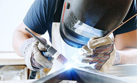 Male in face mask, protective gloves welds with argon-arc welding. Welder makes weld seam on metal frame. Worker dressed in t-shirt and overalls.