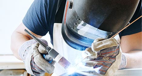 Male in face mask, protective gloves welds with argon-arc welding. Welder makes weld seam on metal frame. Worker dressed in t-shirt and overalls.