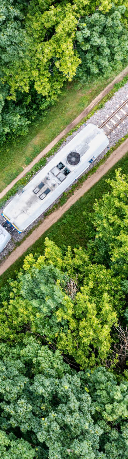 A diesel locomotive pulls the carriages of a passenger train along a railway line through a green deciduous forest. Passenger rail transportation. View from above.